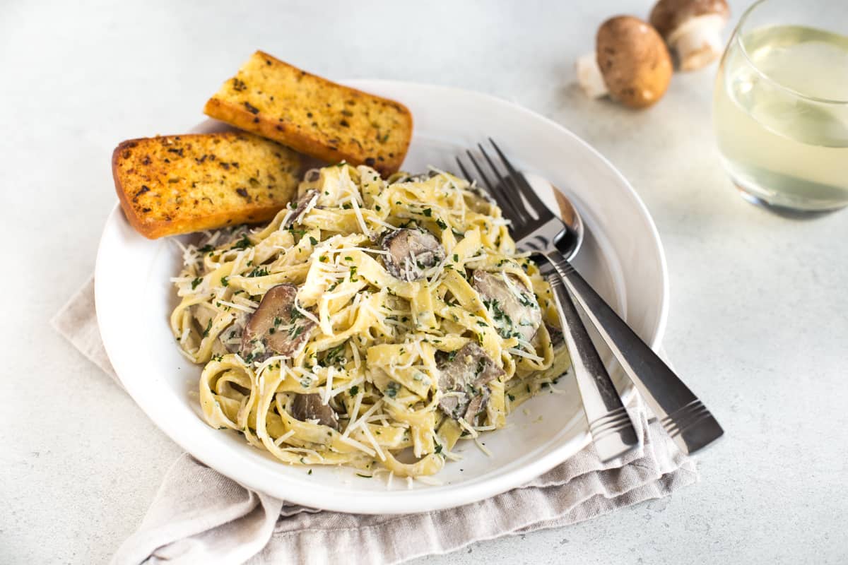 Portion of creamy garlic mushroom tagliatelle in a white bowl with garlic bread.