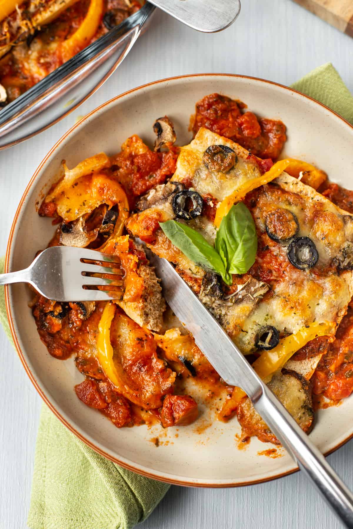 A knife and fork cutting a piece of tofu baked with tomato sauce and cheese.