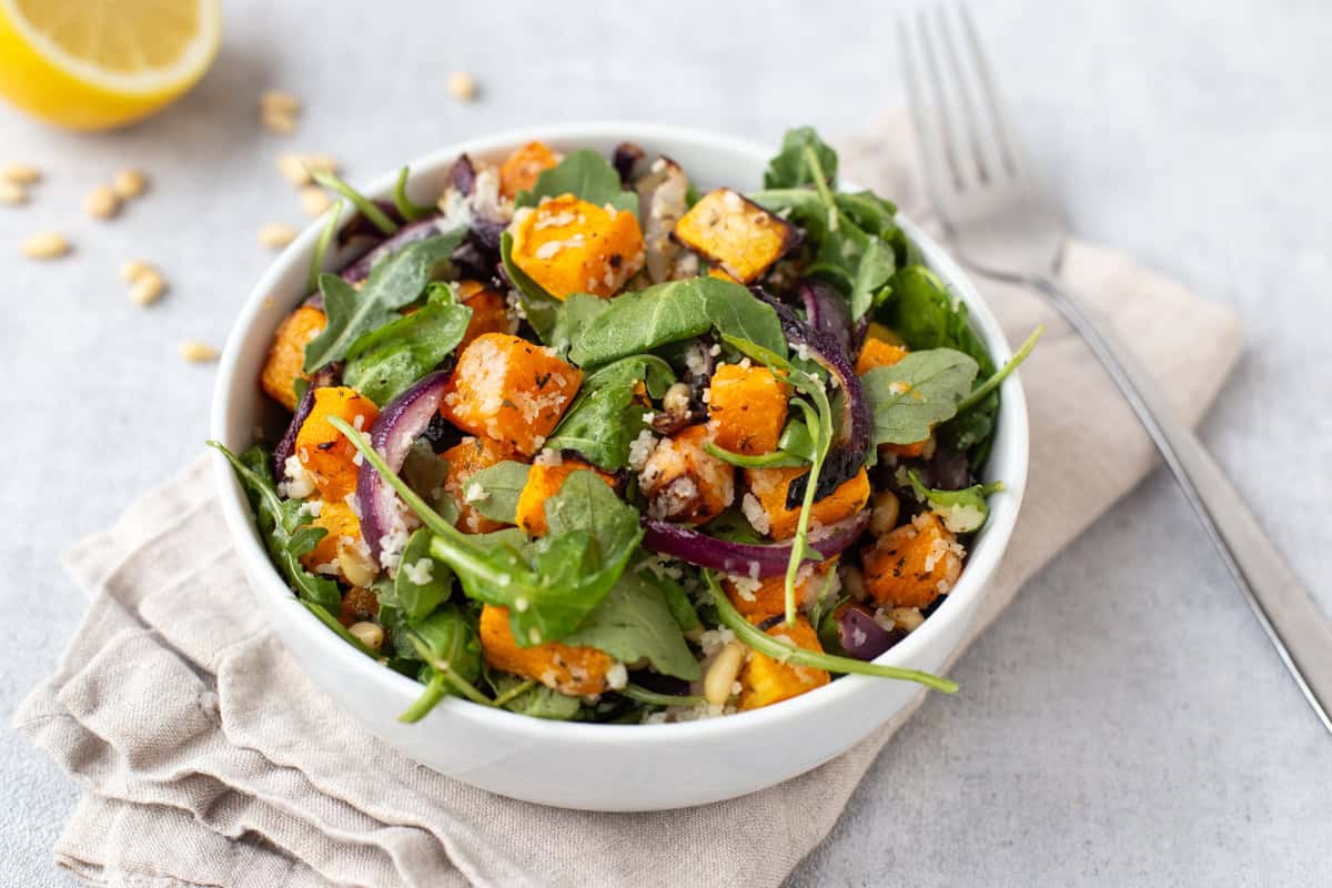 A portion of roasted butternut squash salad in a bowl with red onion and arugula and goat cheese.