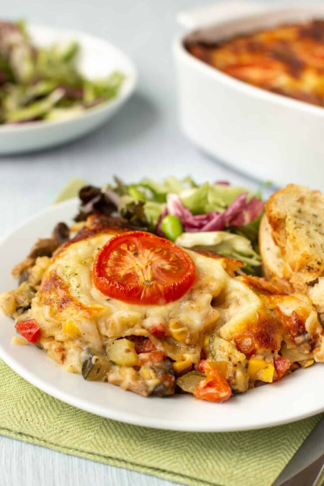 A portion of cheesy vegetable lasagne on a plate with salad and garlic bread.